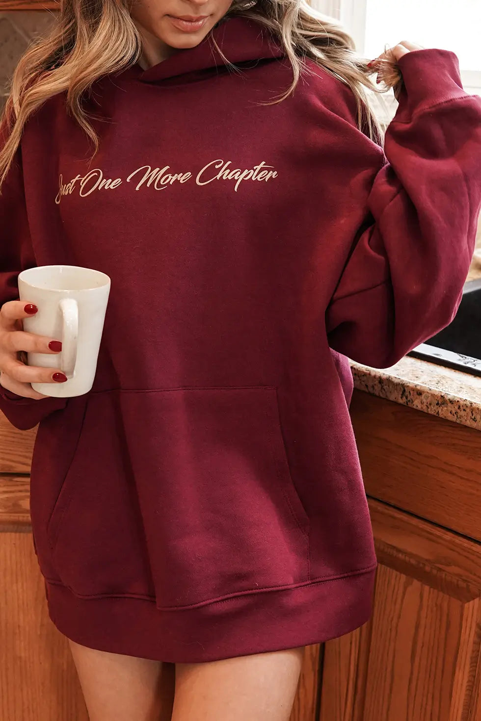 A woman wearing a maroon hoodie with the words "Just One More Chapter" in a script font across the chest area.