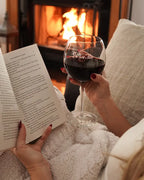 A woman reading a book and holding a glass of red wine in the Pinot wine glass in front of a fireplace.