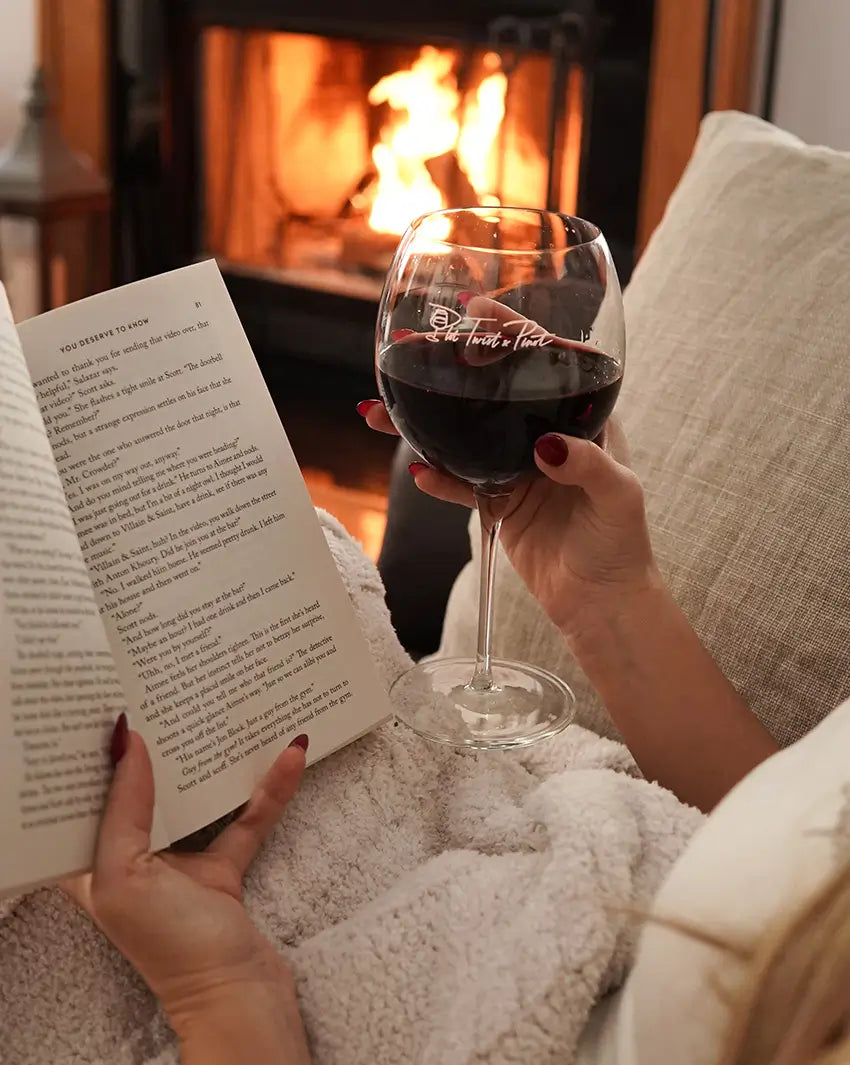 A woman reading a book and holding a glass of red wine in the Pinot wine glass in front of a fireplace.