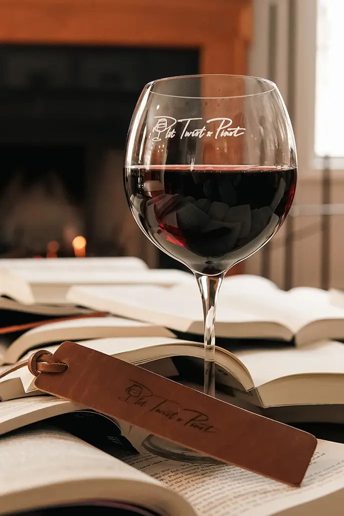 A glass of red wine with script on it is placed on open books alongside a brown leather bookmark. A fireplace with a gentle flame is blurred in the background, creating a cozy atmosphere.