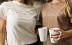 Two people wearing the t-shirts in vintage white and vintage brown colors holding mugs.